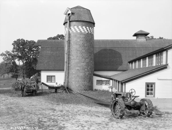 View across farmyard towards a man using an ensilage cutter to fill a silo next to a barn. A Farmall tractor is belt-driving the cutter, and two men are working on a wagon near the silo to feed cornstalks into the cutter.
