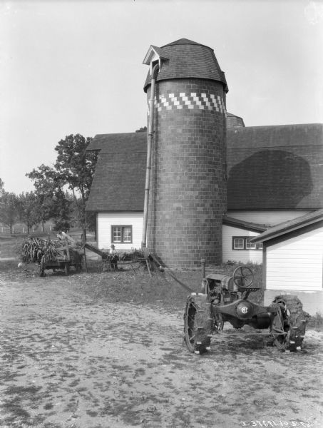 View across farmyard towards a man using an ensilage cutter to fill a silo next to a barn. A Farmall tractor is belt-driving the cutter, and a man is working on a wagon near the silo to feed cornstalks into the cutter.
