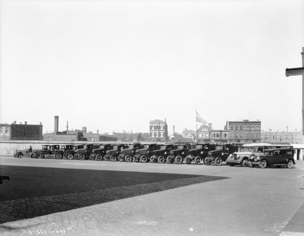 View across factory yard towards a fleet of trucks. On the far left is a man sitting in an automobile, and on the far right a man is sitting in the driver's seat of a harvester coach, and a man is sitting in the driver's seat of an automobile. There are buildings behind the solid wood fence in the background.