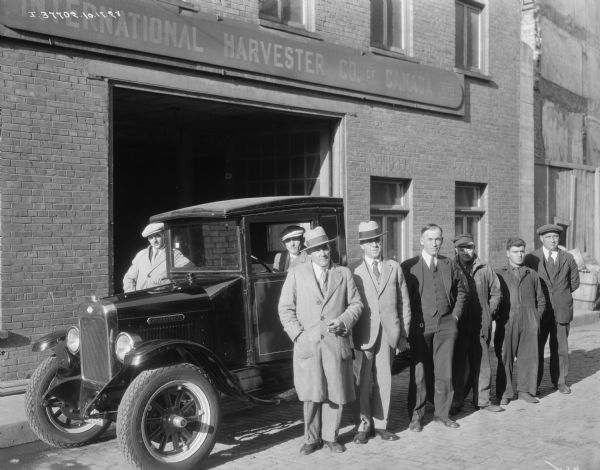 A group of men are standing around an International truck parked in front of an open garage door in a brick building. Some of the men are wearing suits and hats, and the other men are wearing work clothes. A man is sitting in the driver's seat of the truck. The sign above the door reads: "International Harvester Co. of Canada." 
