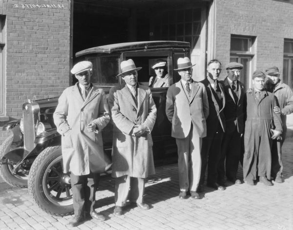 A group of men are standing around an International truck parked in front of an open garage door in a brick building. Some of the men are wearing suits and hats, and the other men are wearing work clothes. A man is sitting in the driver's seat of the truck.