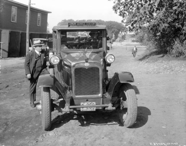 View of front of truck, with a man standing alongside the running board of the passenger side of the International truck which has a Quebec license plate. Another man is sitting in the driver's seat of the truck, which has a sign on the roof that reads: "Simon Service Station, Hull, SH.5169." A group of five small United States and United Kingdom flags are attached to the front of the truck hood, above the Triple Diamond International logo. There are buildings and a road in the background.