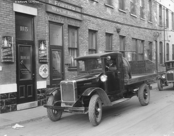 View across street towards a man sitting in the driver's seat of a truck parked alongside a brick building. There is a Quebec license plate on the front of the truck. The sign on the glass doors of the building read: "Bureau, Elz Fortier Ltee Manuf. en Entrepot Licence No. 15." Signs on the brick walls between the doors are for "Ginger Ale Soda Water"and "Frontenac Beer."