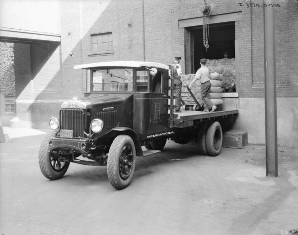 View towards two men standing on the back of a truck which is backed up to a loading dock. They are loading or unloading bales of what may be manila.