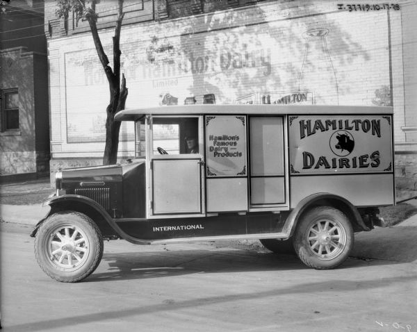View across street towards a man sitting in the driver's seat of a Hamilton Dairies truck parked in front of a brick building. A sign painted on the brick wall of the buildings reads: "Hamilton Dairy."