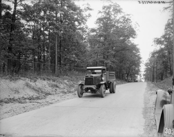 View from side of road towards a man driving an International dump truck. The truck has the Triple Triangle logo on the front. In the background is a fence and trees. In the right foreground is an automobile with an Indiana license plate.