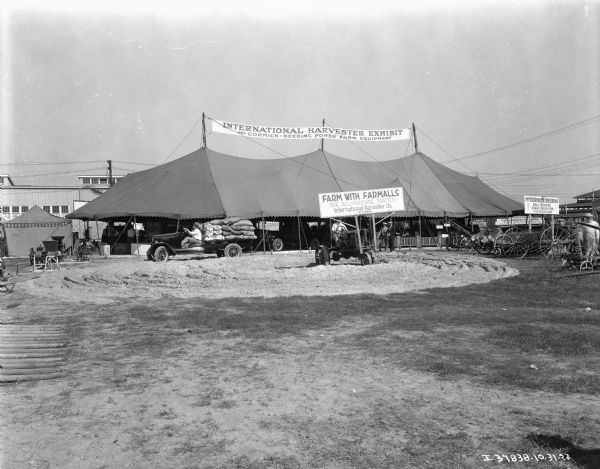 View across fairgrounds towards an International Harvester exhibit tent. The open-sided tent has a man driving a truck loaded heavily with sandbags, a man driving a tractor, and other agricultural implements on display.