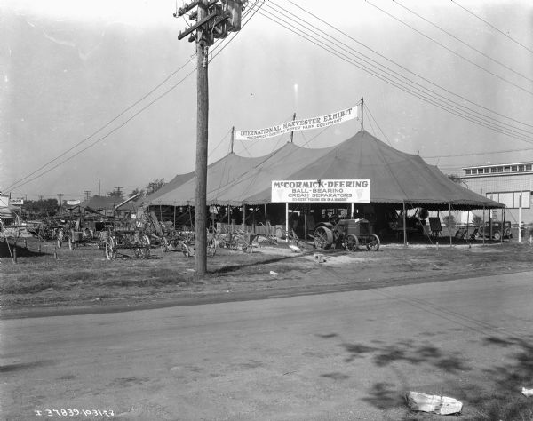 View across fairgrounds towards an International Harvester exhibit tent. The open-sided tent has a tractor and other agricultural implements on display.