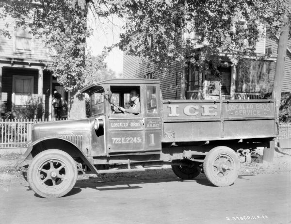 A man is sitting in the driver's seat of an Interanational truck used by the Loscalzo Bros. ice delivery truck parked in a neighborhood.