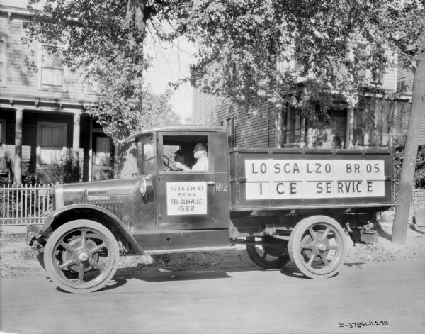 A man is sitting in the driver's seat of an Interanational truck used by the Loscalzo Bros. ice delivery truck parked in a neighborhood.