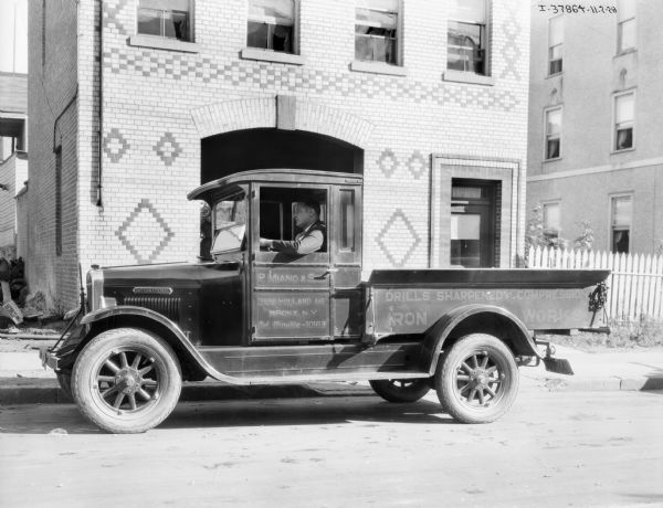 View across street towards a man sitting in the driver's seat of an International truck used by P. Milano. The sign painted on the side of the truck reads: "3556 Holland Ave., Bronrx, N.Y., Drills Sharpened, Compression, Iron Work." The building behind the truck has decorative brickwork, and a large, arched entrance.