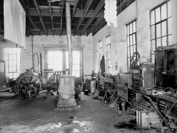 Interior view of a dealership. There is a woodburning stove in the middle of the work space, and a tractor and an automobile are near the back of the room near large windows. Tools and a workbench are along the right wall.