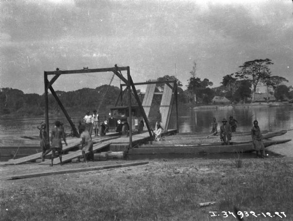 View towards shoreline of people unloading at waterfront in African village. The truck is on a platform supported by four long boats. The far shoreline is in the background.