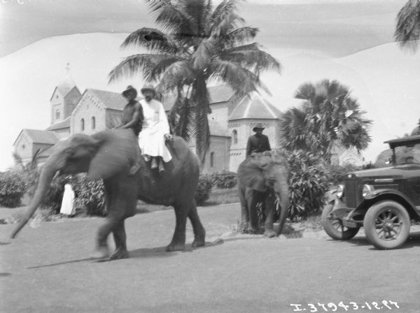 Man are riding two elephants in the yard of large, tropical estate. On the right is an International truck. In the background is a large building.