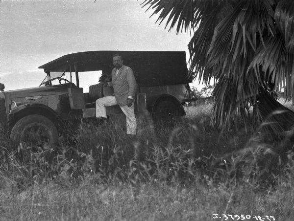 View of a man posing with an International truck in the grass under a coconut tree.