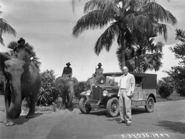 A man is standing in front of a "Sahara" truck. On the left are men sitting on two elephants. Coconut trees are in the background.