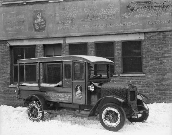 View towards a man sitting in the driver's seat of a delivery truck parked in front of a brick building. The hood of the truck is wrapped with a quilted cover, with an opening for the radiator grille. The sign on the building and on the truck reads: "La Belle Fermiere, Noe Bourassa Limitee." Snow is on the ground.