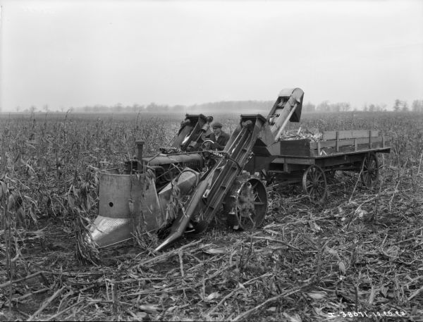 Slightly elevated view of a man driving a Farmall tractor with a McCormick-Deering corn picker and a wagon. The corn picker may be an experimental machine.