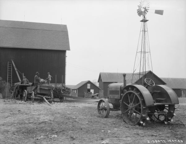 A tractor in the foreground is belt-driving an ensilage cutter near a barn. Men are working on wagons to feed the cutter. There is a windmill behind the tractor on the right near more farm buildings.