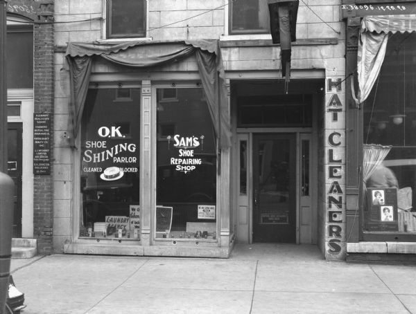 View across sidewalk towards show windows of a shoe repair shop. The sign on the windows reads: "O.K. Shoe Shining Parlor" and "Sam's Shoe Repairing Shop." The reflection of the photographer standing with his camera on a tripod is in the window of the door.