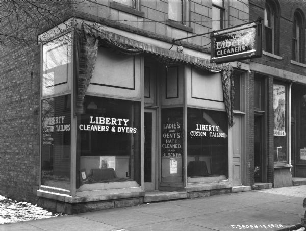 View of storefront for Liberty Cleaners & Dyers. The awning is folded back, and a sign printed on the side of the awning reads: "Pressing While U Wait." A sign on a window near the door reads: "Ladie's and Gent's Hats Cleaned and Blocked." Snow is on the ground next to the side of the building on the left.