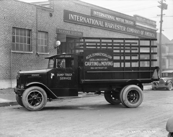 View across street towards a truck with a sign on the driver's side door that reads: "Dump Truck Service." The sign on the stake body of the truck bed reads: "Alex Zembrzycki & Sons, Local & Long Distance Carting & Moving." The signs on the brick building in the background reads: "International Motor Trucks" and "Service Station."