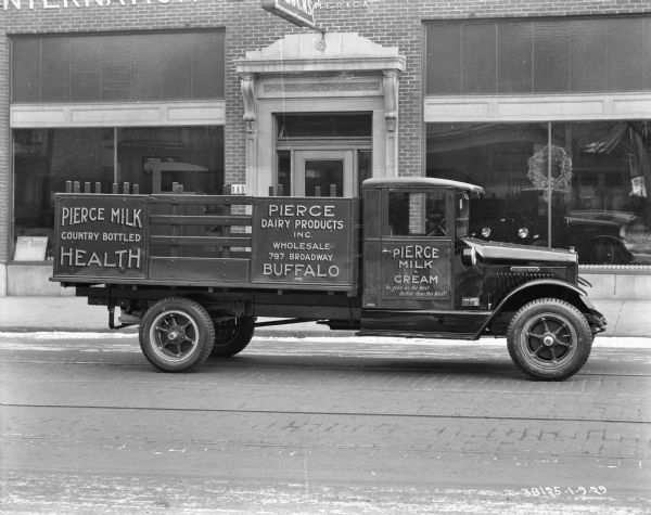 View across street towards a truck parked near the curb in front of a brick building with show windows. The signs painted on the side of the truck reads: "Pierce Milk Country Bottled Health" and "Pierce Milk & Cream." Trucks are on display inside the building.