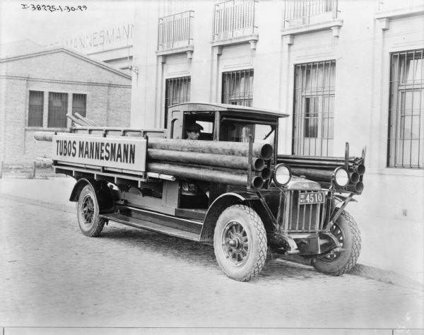 View across street towards a man sitting in the cab of a truck parked along a curb in front of a large building. The sign on the truck reads: "Tubos Mannesmann." Long pipes are on racks that run the length of the truck. More pipes are stacked in the truck bed.