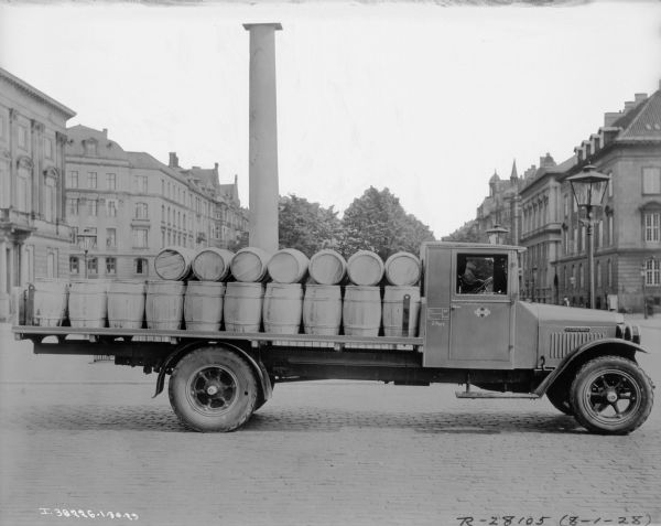 Right side profile view of a man sitting in the driver's seat of a truck parked on a cobblestone street. The open bed of the truck is stacked with barrels. The printing on the barrels reads: "Danish Butter." In the background is a column, trees, and buildings along both sides of the street.