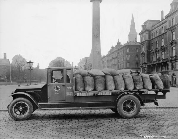 View of a man sitting in the driver's seat of an International truck parked on a cobblestone street in a town. A sign on the driver's side door reads: "Hollose Molle." Sacks are stacked on the open truck bed. There is a column and trees behind the truck, and buildings line both sides of the boulevard in the background.