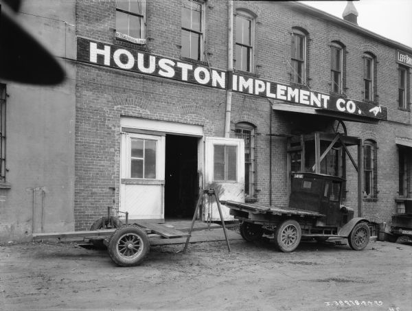 A truck with trailer and a hoist for transporting Farmalls is parked in front of a building with a sign for "Houston Implement." There is a sign in a second story window for "The Texas Artificial Limb Co." and on the far right is a sign for the "Leff Bros."
