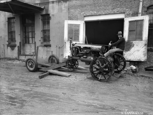 A man is driving a tractor onto a wagon just outside the open double doors of a brick building at a dealership.