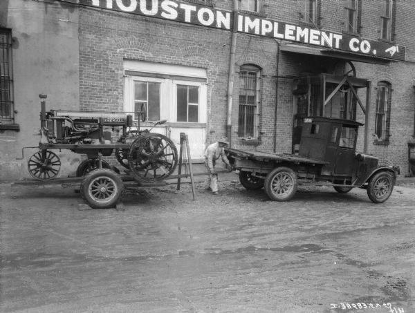 A ma is standing near a truck with trailer and a hoist for transporting Farmalls is parked in front of a building with a sign for "Houston Implement."