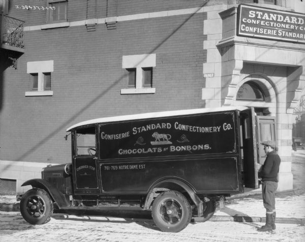 A man is standing at the open rear door or a Confiserie Standard Confectionery truck. The truck is parked along the curb near a brick building on a street corner. The sign above the door reads: "Standard Confectionery Co." Snow is on the ground.