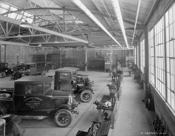 Elevated view of a large room at a dealership. The ceiling rafters are exposed, and there are skylights. Men are working at work tables on the right, and trucks are parked on the left.
