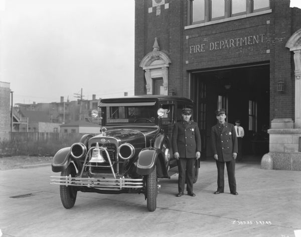 Two firefighters are standing next to a fire department ambulance parked outside the open doorway of a Fire Department building. Another man is standing inside the station.