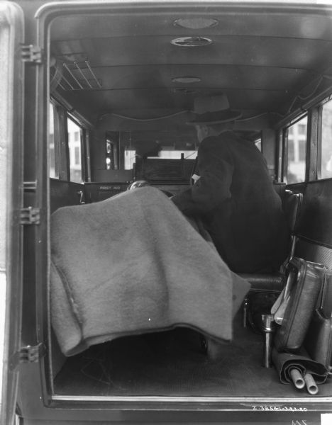 View from rear of ambulance with open door towards a man sitting with another man laying under a blanket on a stretcher.