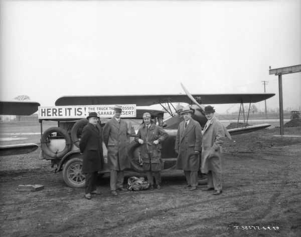 Five men are standing in front of a truck with a sign on it that reads: "Here It Is! The Truck That Crossed the Sahara Desert." Airplanes are parked behind the truck.
