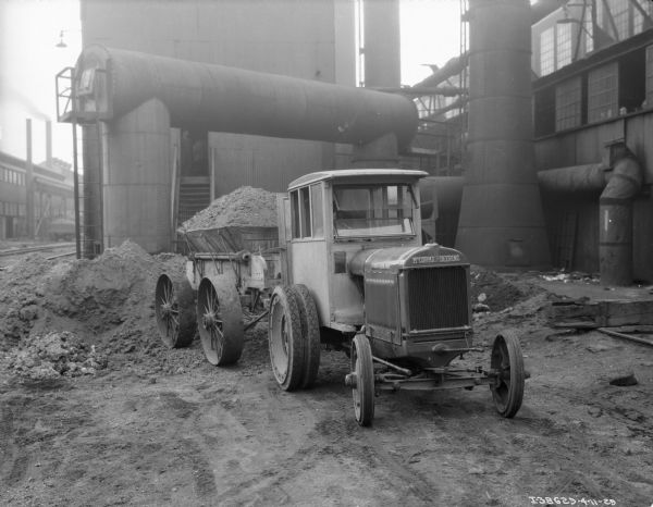 View towards the front of a McCormick-Deering truck pulling a loaded wagon. In the background are industrial buildings.