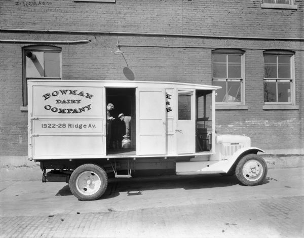 Right side profile view of a parked Bowman Dairy delivery truck. The side doors of the truck are open, and two men are seen through the open doors while working what may be a loading dock on a large brick building behind the truck.