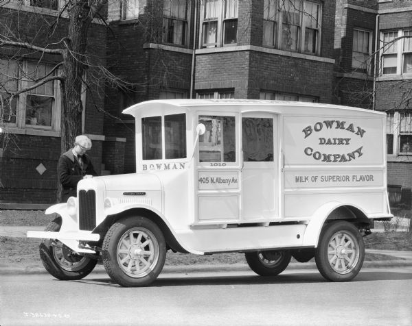 View across street towards a Bowman Dairy delivery truck parked along a curb. A sign painted on the side of the truck reads: "Milk of Superior Flavor." A man is standing behind the truck. There are brick buildings in the background.