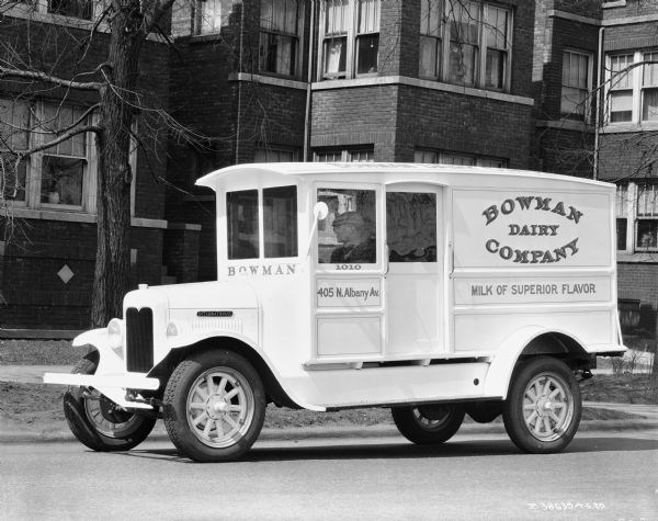 View across street towards a man sitting in the driver's seat of a Bowman Dairy delivery truck. A sign painted on the side of the truck reads: "Milk of Superior Flavor." Brick buildings are in the background.