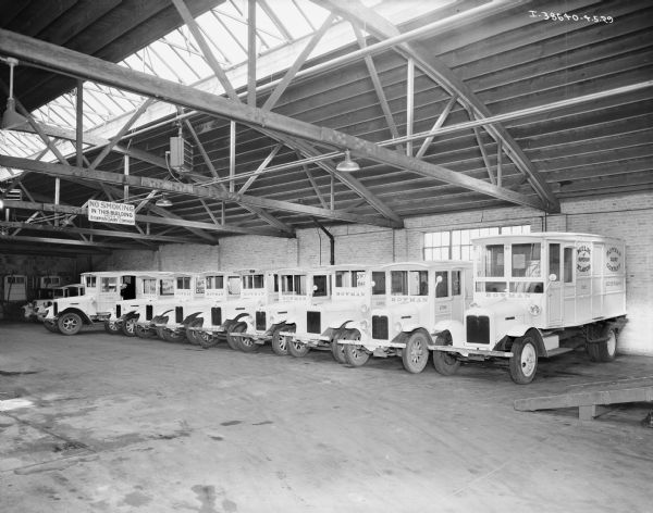 View of fleet of trucks parked indoors in a large garage with a skylight. There is a man in the driver's seat of a truck in the background on the left.