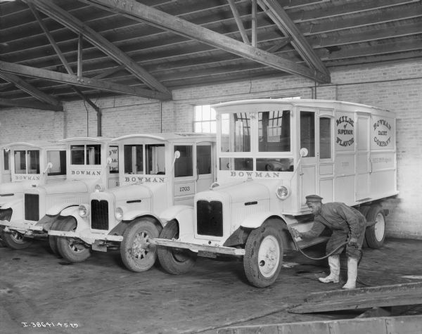 A man is standing on the far right next to a Bowman Dairy Company delivery truck in a brick building with an exposed ceiling. The man is wearing a hat, coveralls, and boots and is holding a spray head attached to a hose. Four other Bowman trucks are parked in a row towards the left.