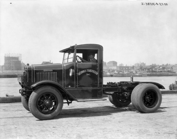 View of a man sitting in the driver's seat of a Greenwich Transportation Inc. truck parked on a pier. In the background is water and a city on the far shoreline. The back section of the truck behind the cab is exposed.