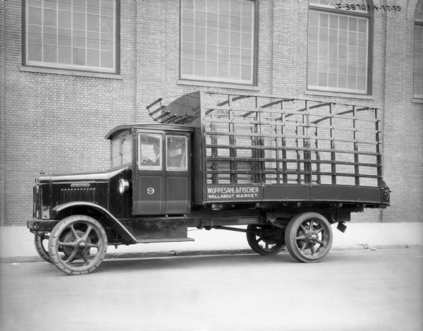 View across street towards a man sitting in the driver's seat of a truck that has a sign on the side of the bed that reads: "Wuppesahl & Fisher, Wallabout Market." There is a brick building in the background.