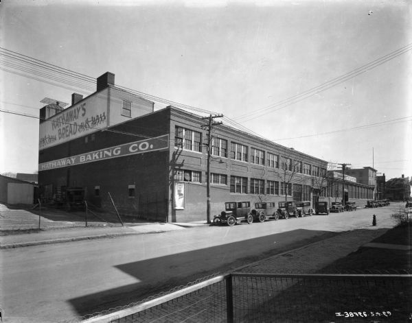 View across street towards automobiles parked along the curb in front of a large brick building. A sign painted on the side of the building reads: "Hathaway's Bread." 