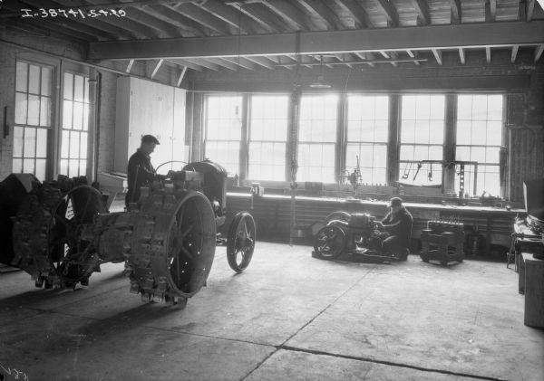 A man is working on a tractor, and another man is working on an engine in a repair shop at a dealership. A row of windows are in the background.