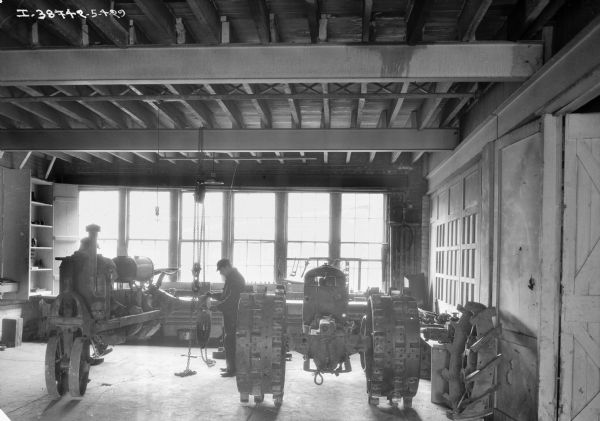 Two men are working on an engine and a tractor in a repair shop at a dealership. A row of windows are in the background.
