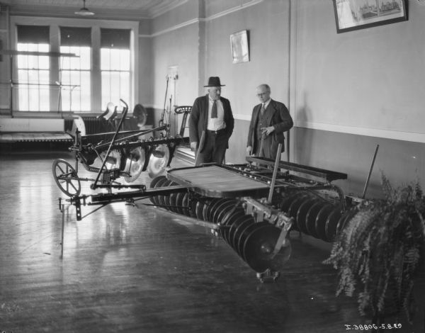 Two men are standing inside a dealership looking at a disk harrow.